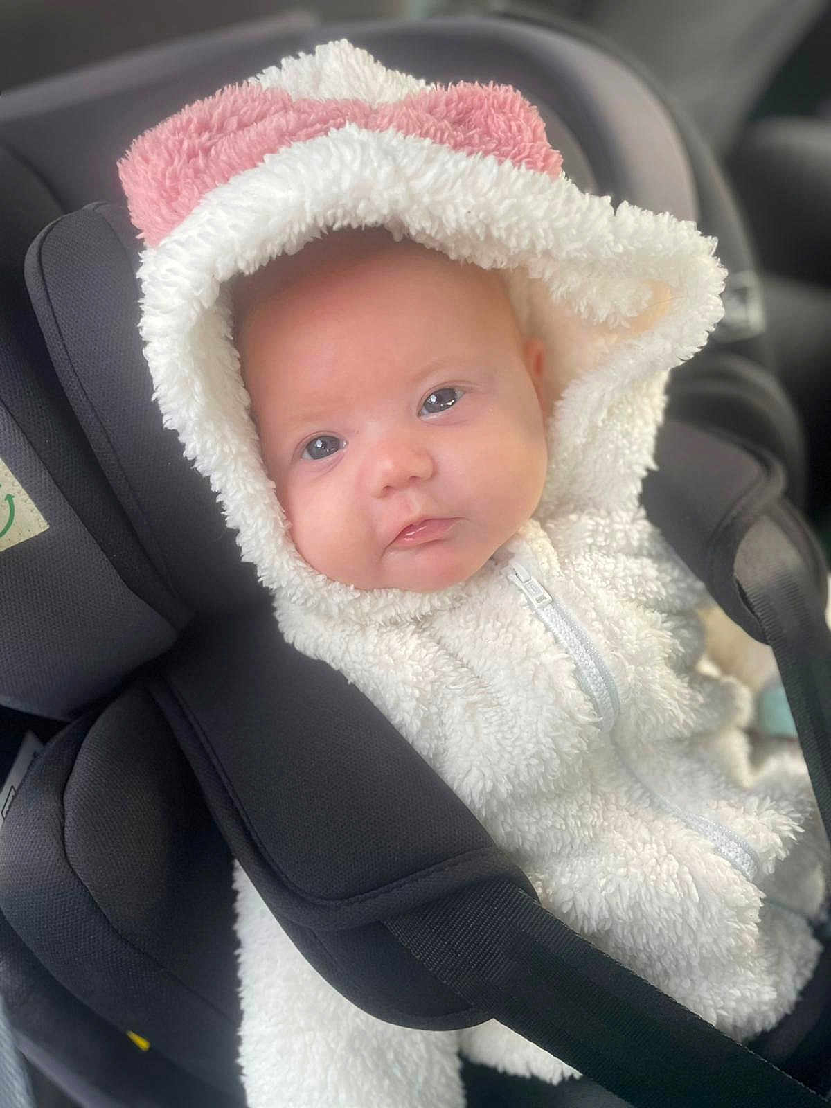 Mali-Rae is registered to the contest to win money with this photo: baby, infant, car_seat, hood, white_jacket, pink_detail, face, eyes, clothing, child, seat_belt, soft_texture, cute, portrait, indoor, person, warm_clothing, closeup, newborn, babywear