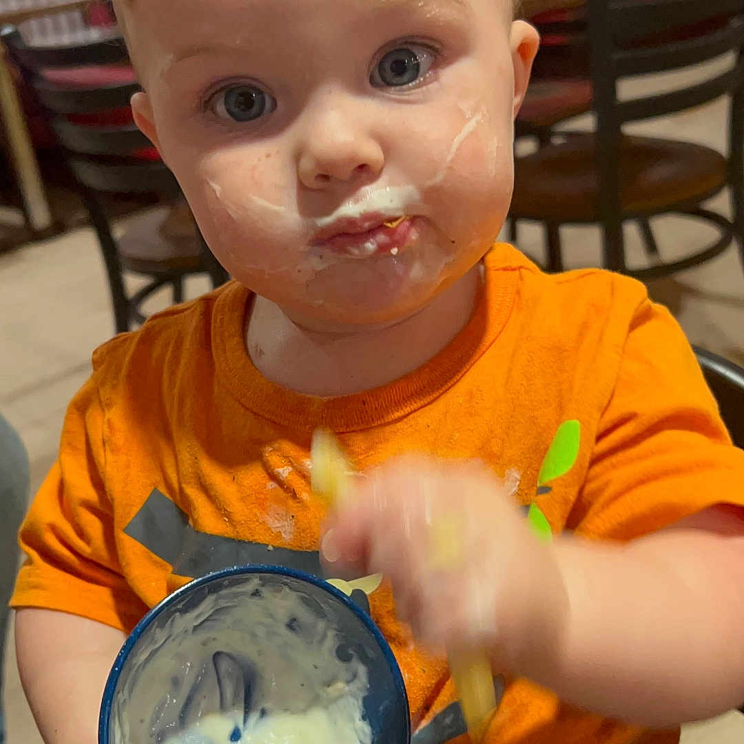 Kayyson is registered to the contest to win money with this photo: blond_hair, blue_cup, casual, chair, child, closeup, curious_expression, cute, dining_area, eating, food, hand_motion, indoor, meal_time, messy_face, orange_shirt, person, portrait, table, toddler