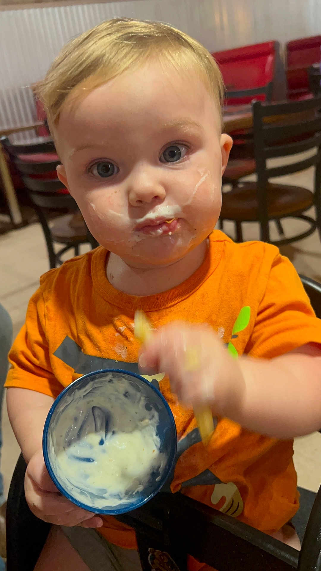 Kayyson is registered to the contest to win money with this photo: toddler, child, messy_face, orange_shirt, blue_cup, food, eating, hand_motion, indoor, dining_area, chair, table, blond_hair, curious_expression, closeup, cute, casual, meal_time, person, portrait