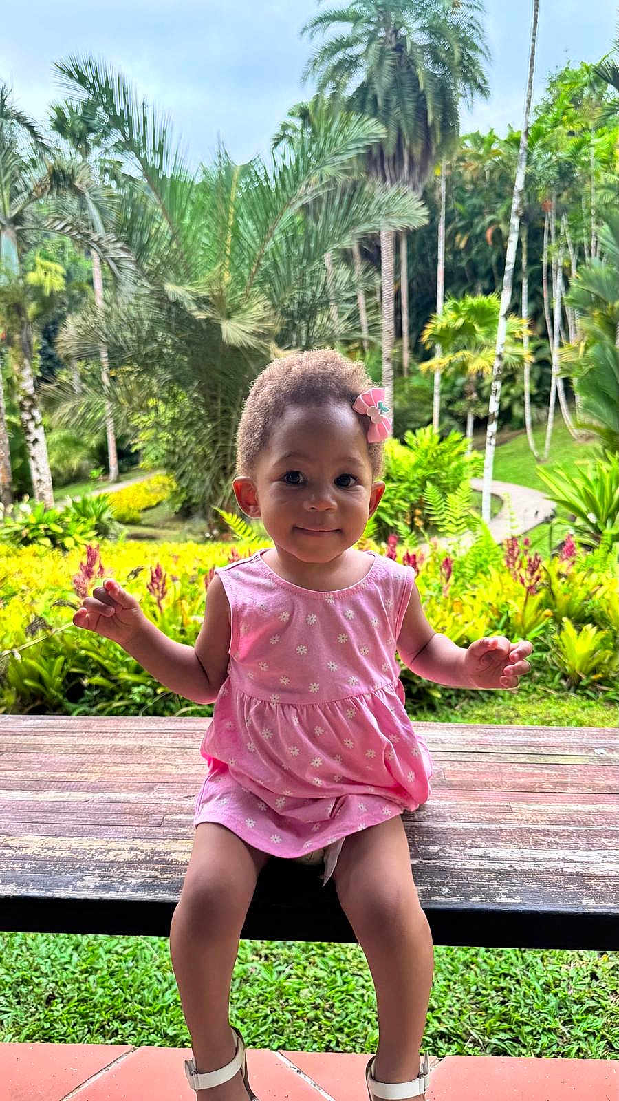 Kehlany a rejoint le concours — aidez-le/la à gagner de superbes lots ! toddler, child, pink_dress, flower_hair_clip, bench, wooden_bench, sandals, greenery, tropical_plants, flowers, outdoor, nature, smiling, portrait, cute, garden, grass, trees, sunlight, happy