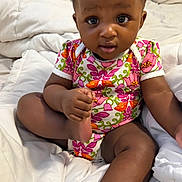Zuri is registered to the contest to win money with this photo: baby, infant, child, onesie, floral_pattern, blanket, bed, comfort, cute, expression, skin, foot, hand, sitting, indoors, person, young, portrait, head, eyes