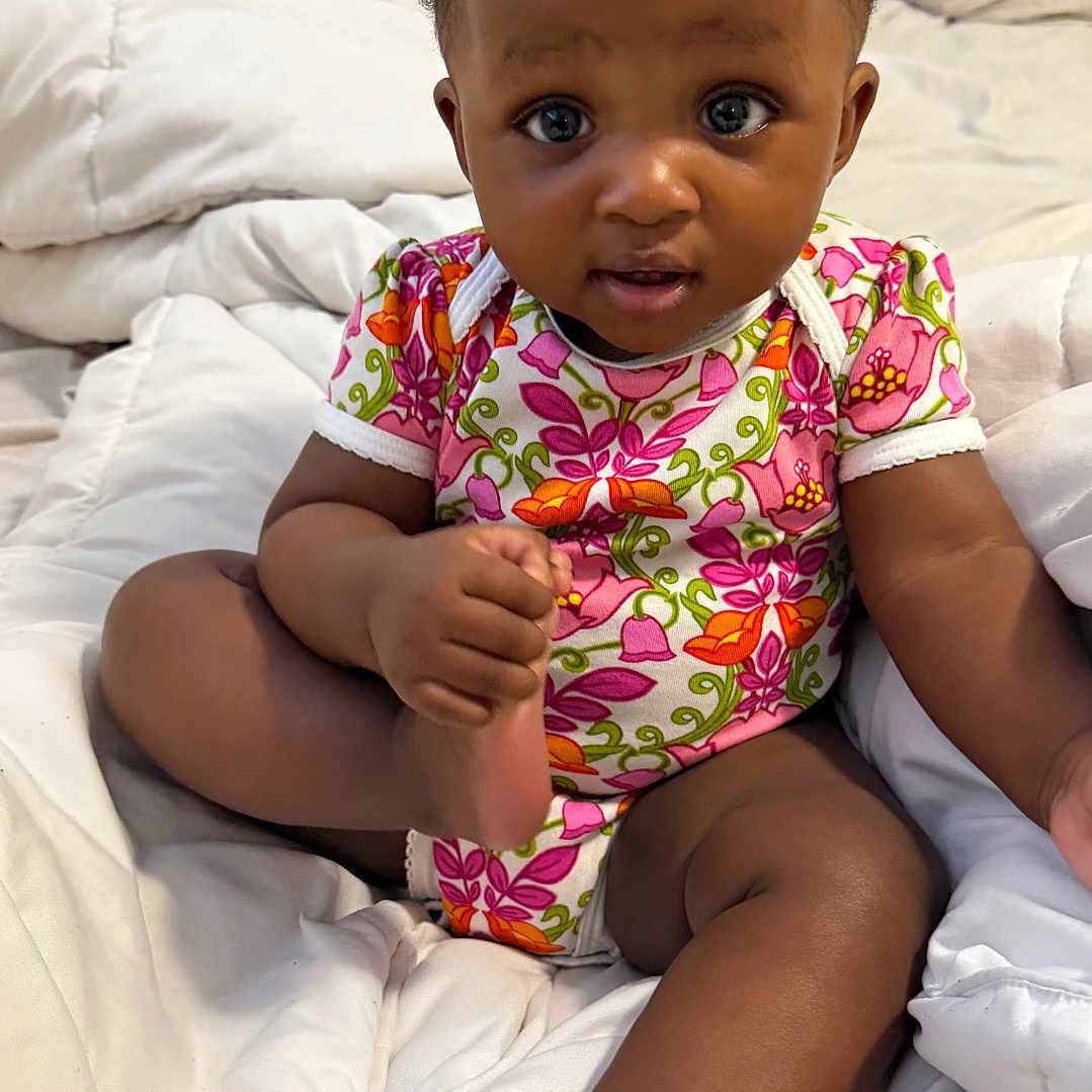 Zuri is registered to the contest to win money with this photo: baby, infant, child, onesie, floral_pattern, blanket, bed, comfort, cute, expression, skin, foot, hand, sitting, indoors, person, young, portrait, head, eyes