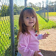 Charlie is registered to the contest to win money with this photo: child, girl, smiling, outdoor, daylight, fence, bench, grass, field, nature, headband, long_hair, casual_clothing, happy, portrait, person, sunlight, park, playground, cute