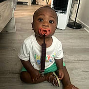 Davonta is registered to the contest to win money with this photo: toddler, child, pacifier, floor, indoor, white_shirt, green_shorts, sitting, curious, baby, footwear, sandals, clip, wooden_floor, person, home, young_child, expression, looking_up, cute