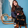 woman, dog, rottweiler, sled, blue_background, indoor, smiling, casual_clothing, long_hair, seated, wooden_furniture, pet, animal, tongue_out, friendship, portrait, studio, bare_branches, happy, leisure