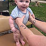 baby, child, person, hands, tattoos, striped_clothing, outdoor, grass, golf_cart, seat, summer, cute, infant, blue_eyes, expression, holding, nature, trees, daylight, foot