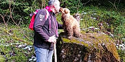 Perle participe au concours pour gagner de l'argent avec cette photo : man, dog, rock, forest, moss, hiking, walking_stick, outdoor, nature, tree, greenery, snow, pet, animal, backpack, curly_hair, interaction, friendship, standing, casual_clothing