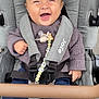 baby, stroller, smiling, sweater, pacifier, clip, seatbelt, child, happy, person, clothing, fabric, footwear, sitting, outdoor, pale_skin, head, hand, toy, comfort
