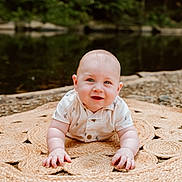 Oliver is registered to the contest to win money with this photo: baby, infant, smiling, outdoor, nature, river, mat, woven_mat, grass, pebbles, cute, child, portrait, face, hands, crawling, shirt, summer, greenery, relaxing