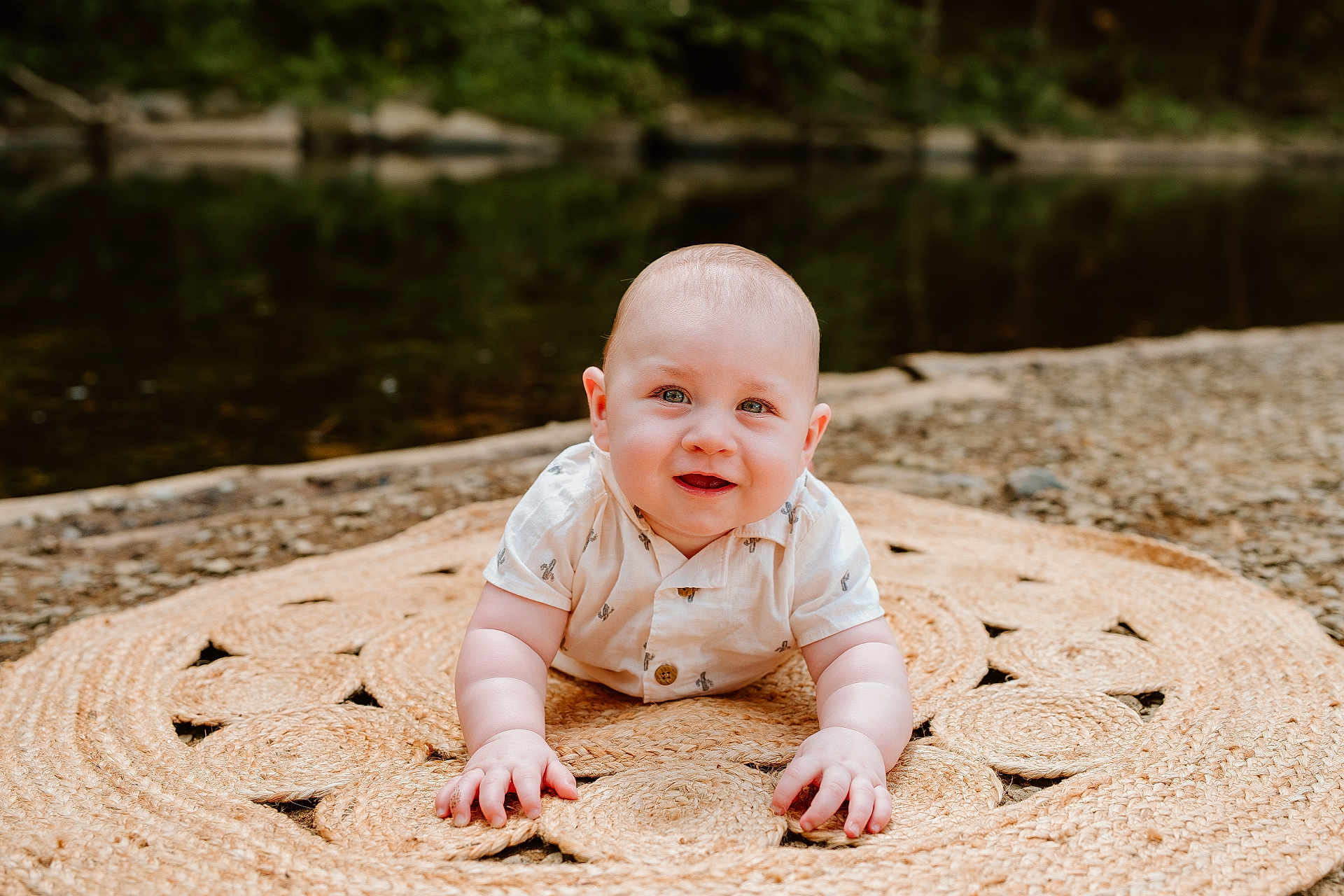 Oliver is registered to the contest to win money with this photo: baby, infant, smiling, outdoor, nature, river, mat, woven_mat, grass, pebbles, cute, child, portrait, face, hands, crawling, shirt, summer, greenery, relaxing