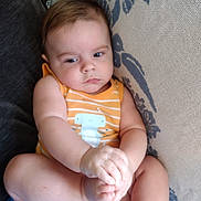 Braxton joined the competition — help win amazing prizes! baby, child, infant, onesie, orange_clothing, striped_clothing, couch, cushion, hands, feet, sitting, indoor, face, skin, expression, curious, calm, person, young, portrait