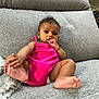 baby, child, infant, pink_clothing, sitting, couch, grey_couch, thumb_in_mouth, barefoot, bracelet, indoor, portrait, cute, curious, relaxed, home, furniture, soft_texture, casual, one_person