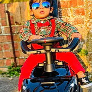 Derrick is registered to the contest to win money with this photo: toddler, child, sunglasses, toy_car, red_suspender, red_bow_tie, plaid_shirt, black_shoes, outdoor, brick_wall, sunlight, reflection, steering_wheel, fashion, portrait, cute, playful, person, casual, ground