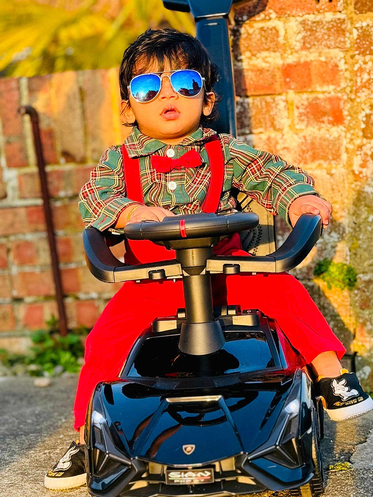 Derrick is registered to the contest to win money with this photo: toddler, child, sunglasses, toy_car, red_suspender, red_bow_tie, plaid_shirt, black_shoes, outdoor, brick_wall, sunlight, reflection, steering_wheel, fashion, portrait, cute, playful, person, casual, ground