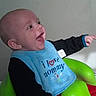 arm, baby, bib, bumbo_seat, cheek, clothing, diaper_organizer, ear, face, green_seat, hand, happy, indoor, infant, laughing, portrait, sitting, smiling, text_on_bib, wall