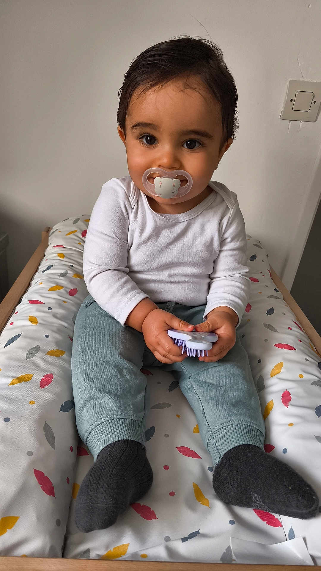 Zakaria a rejoint le concours — aidez-le/la à gagner de superbes lots ! toddler, child, pacifier, brush, sitting, cushion, feather_pattern, white_shirt, blue_pants, dark_socks, indoor, portrait, cute, baby, furniture, wooden_frame, wall_socket, curly_hair, looking_at_camera, hands