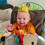 baby, child, crown, orange_shirt, smiling, happy, toy, plush_toy, indoor, chair, blanket, face, hand, cute, infant, sitting, blue_eyes, wall, home, cozy