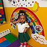 afro_hairstyle, bow, child, colorful, educational_toy, foam_floor, girl, green_skirt, indoor, interactive_wall, peace_sign, playroom, portrait, rainbow_wall, school_uniform, sneakers, striped_socks, toddler, wall_art, white_shirt