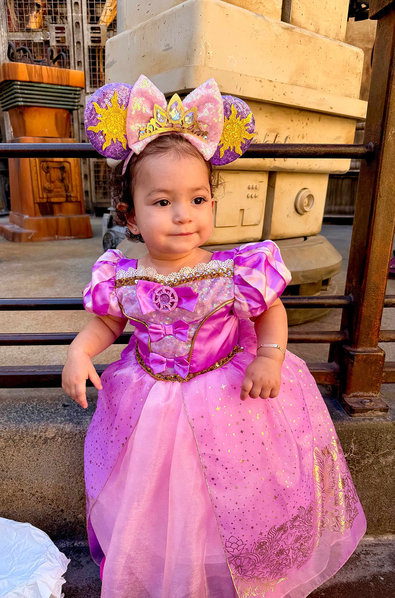 Madeline joined the competition — help win amazing prizes! child, toddler, princess_dress, pink_dress, mouse_ears, headband, sparkles, cute, portrait, outdoor, curly_hair, bracelet, seated, expression, metal_railings, industrial_background, young_child, costume, fashion, person