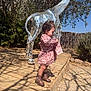 Madeline joined the competition — help win amazing prizes! child, toddler, pink_dress, brown_boots, curly_hair, mosaic_giraffe, sculpture, wooden_platform, outdoor, sunny, shadow, plants, trees, nature, daylight, playful, curious, side_view, fence, gravel