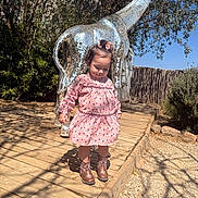 Madeline is registered to the contest to win money with this photo: toddler, child, girl, pink_dress, cowboy_boots, mosaic_giraffe, sculpture, outdoor, sunny, wooden_path, plants, trees, shadows, curly_hair, hair_bow, nature, daylight, playful, cute, portrait