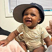 Quentin is registered to the contest to win money with this photo: baby, child, smiling, hat, bucket_hat, onesie, nike, indoor, window, blinds, blanket, bottle, sitting, happy, cute, infant, person, portrait, skin, clothing