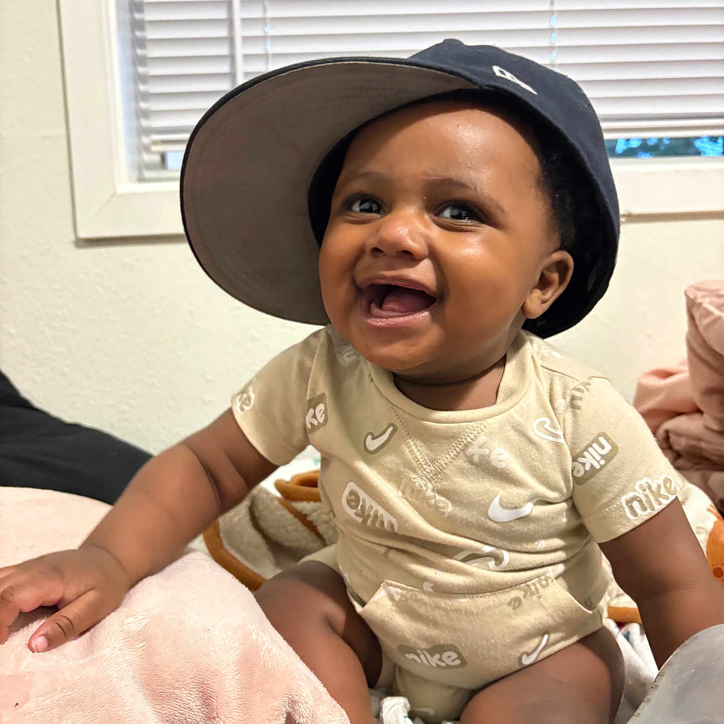 Quentin is registered to the contest to win money with this photo: baby, child, smiling, hat, bucket_hat, onesie, nike, indoor, window, blinds, blanket, bottle, sitting, happy, cute, infant, person, portrait, skin, clothing