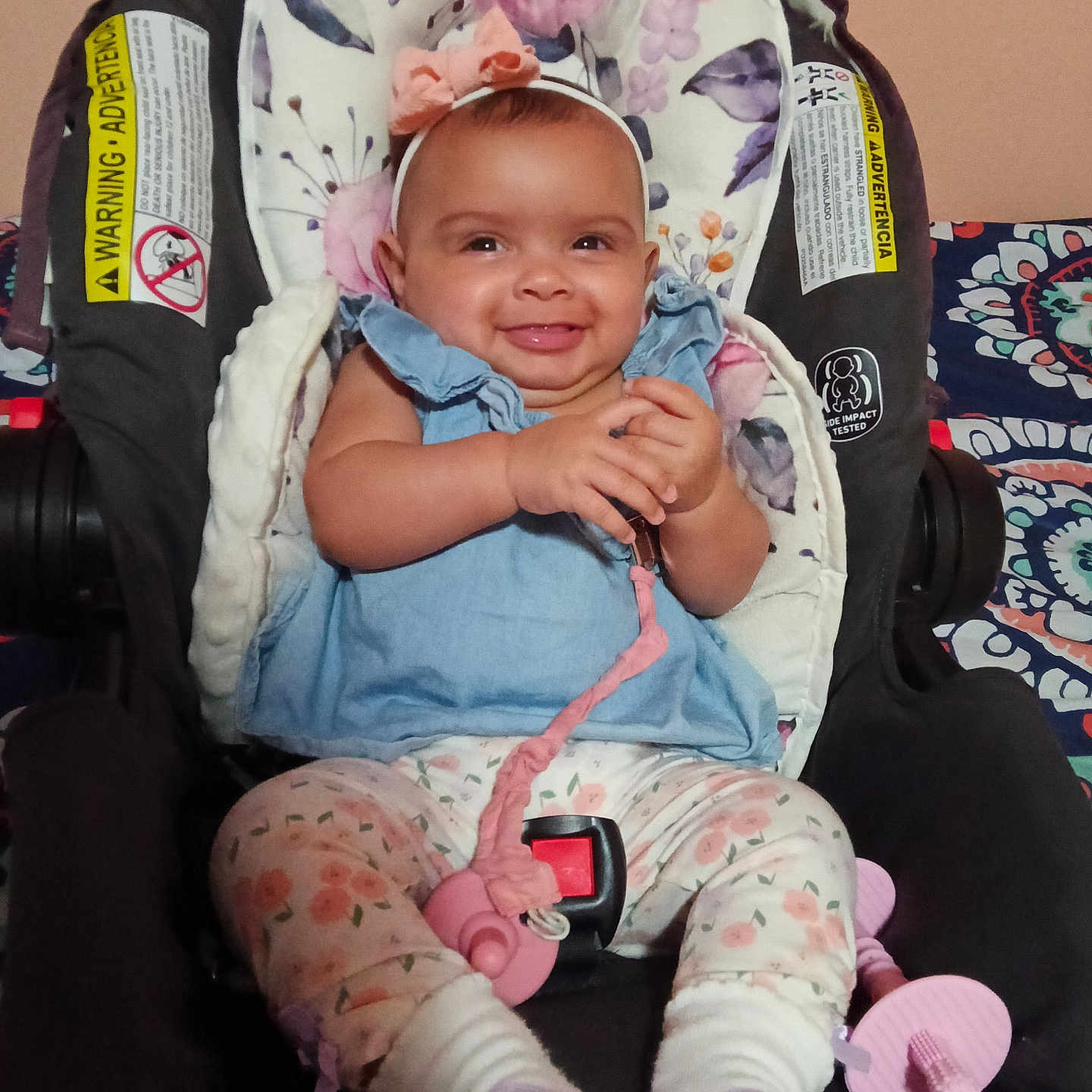 Kodiana is registered to the contest to win money with this photo: baby, infant, child, smiling, car_seat, harness, buckled, floral, headband, blue_dress, pink_pacifier, patterned_clothing, socks, seated, portrait, indoors, vehicle_safety, carseat, happy, closeup