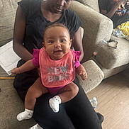 Jahmina is registered to the contest to win money with this photo: baby, woman, couch, living_room, smiling, bib, pink, black_clothing, person, floor, water_bottle, paper, socks, indoor, seated, happy, child, casual, relaxed, family