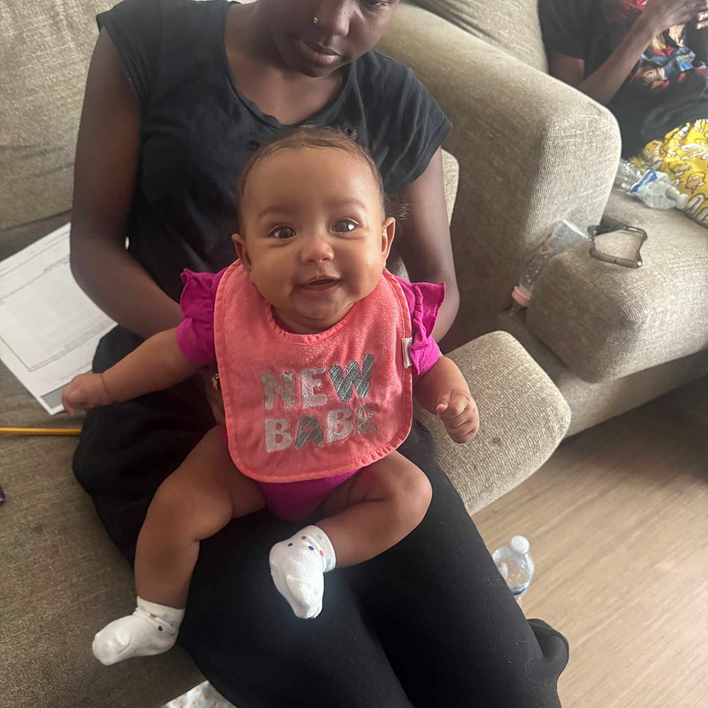Jahmina is registered to the contest to win money with this photo: baby, bib, black_clothing, casual, child, couch, family, floor, happy, indoor, living_room, paper, person, pink, relaxed, seated, smiling, socks, water_bottle, woman