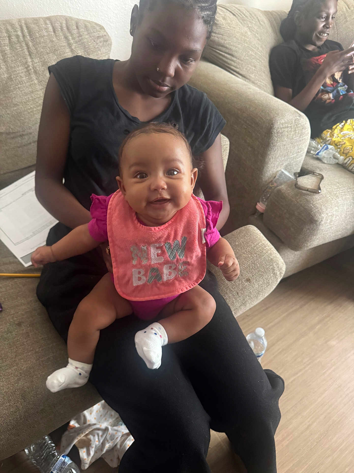 Jahmina is registered to the contest to win money with this photo: baby, woman, couch, living_room, smiling, bib, pink, black_clothing, person, floor, water_bottle, paper, socks, indoor, seated, happy, child, casual, relaxed, family