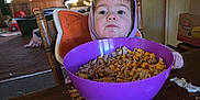 Samuel is registered to the contest to win money with this photo: toddler, child, cereal, purple_bowl, table, wooden_chair, hoodie, indoor, living_room, deer_mounts, family_pictures, snack_time, food, messy_table, window, curtains, cardboard_box, popcorn, face, person