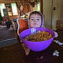 cardboard_box, cereal, child, curtains, deer_mounts, face, family_pictures, food, hoodie, indoor, living_room, messy_table, person, popcorn, purple_bowl, snack_time, table, toddler, window, wooden_chair