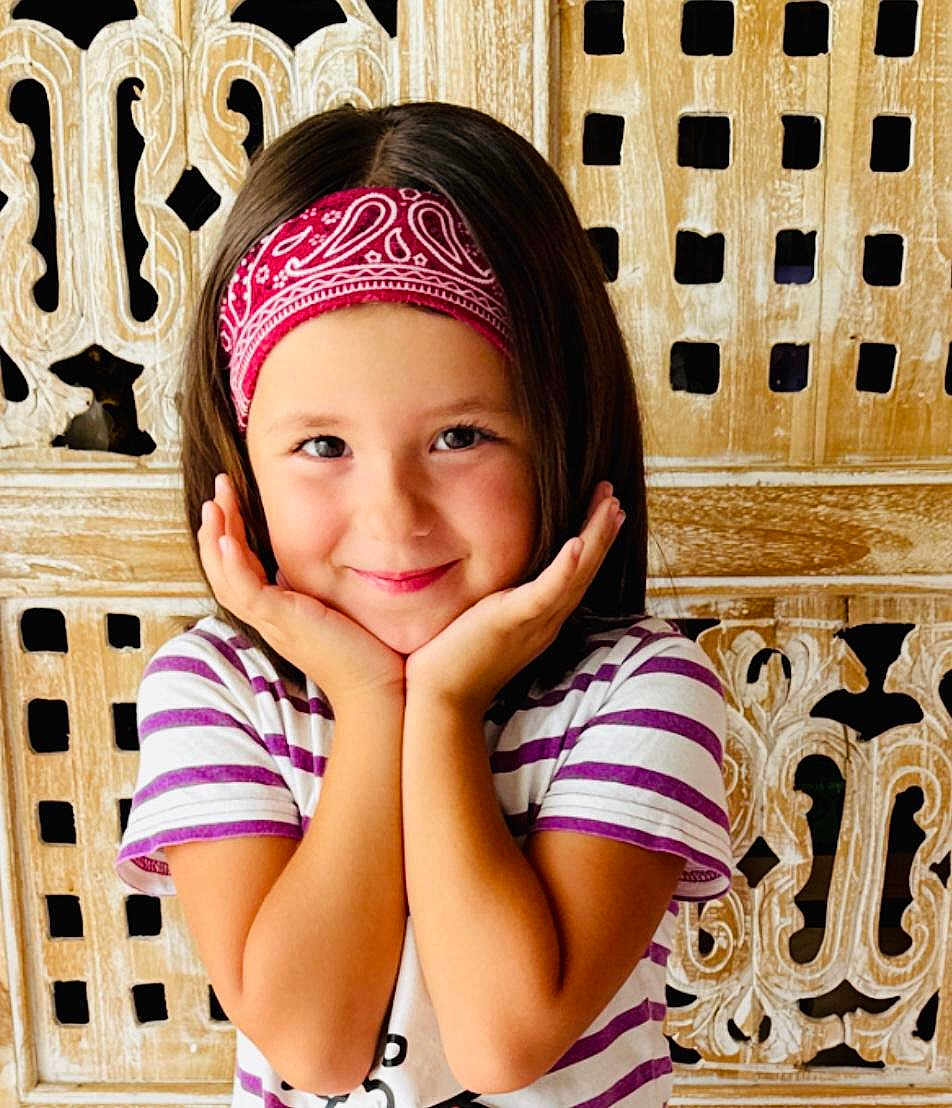 Milla a rejoint le concours — aidez-le/la à gagner de superbes lots ! child, smiling, bandana, striped_shirt, portrait, happy, cute, hands_on_face, wooden_background, short_hair, indoors, person, face, young, playful, posing, casual_clothing, headband, childhood, bright