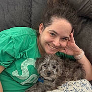 Max is registered to the contest to win money with this photo: woman, person, dog, puppy, pet, portrait, smiling, happy, indoors, couch, blanket, green_shirt, wristwatch, phone, dark_couch, home, cuddle, seated, gray_fur, photography