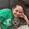 woman, person, dog, puppy, pet, portrait, smiling, happy, indoors, couch, blanket, green_shirt, wristwatch, phone, dark_couch, home, cuddle, seated, gray_fur, photography
