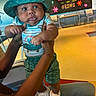 Andrez joined the competition — help win amazing prizes! baby, child, hat, dinosaur, striped_shirt, shorts, sneakers, hands, indoor, bulletin_board, flowers, text, colorful, seated_adult, curious_expression, yellow_floor, chairs, table, person, well_lit