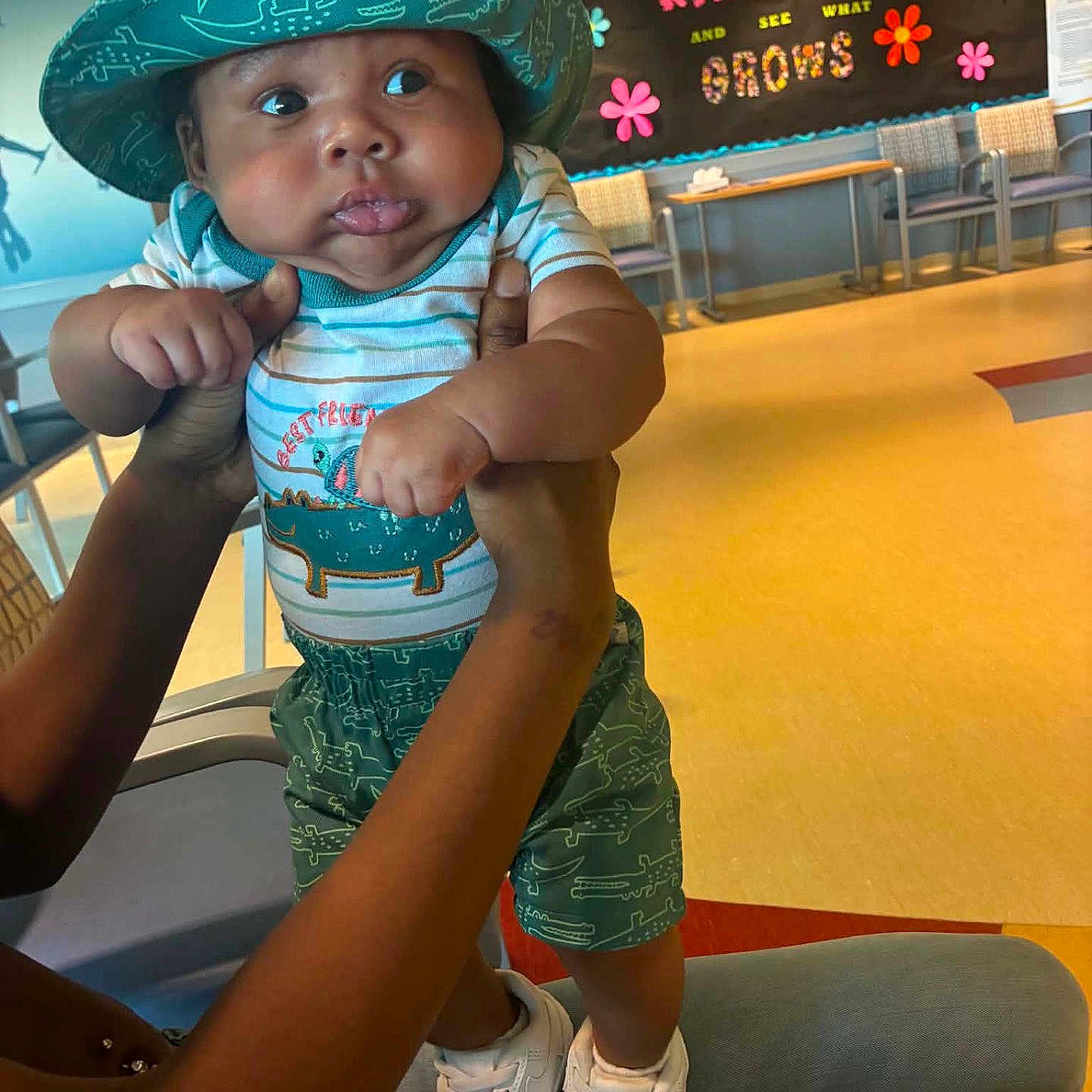 Andrez joined the competition — help win amazing prizes! baby, child, hat, dinosaur, striped_shirt, shorts, sneakers, hands, indoor, bulletin_board, flowers, text, colorful, seated_adult, curious_expression, yellow_floor, chairs, table, person, well_lit