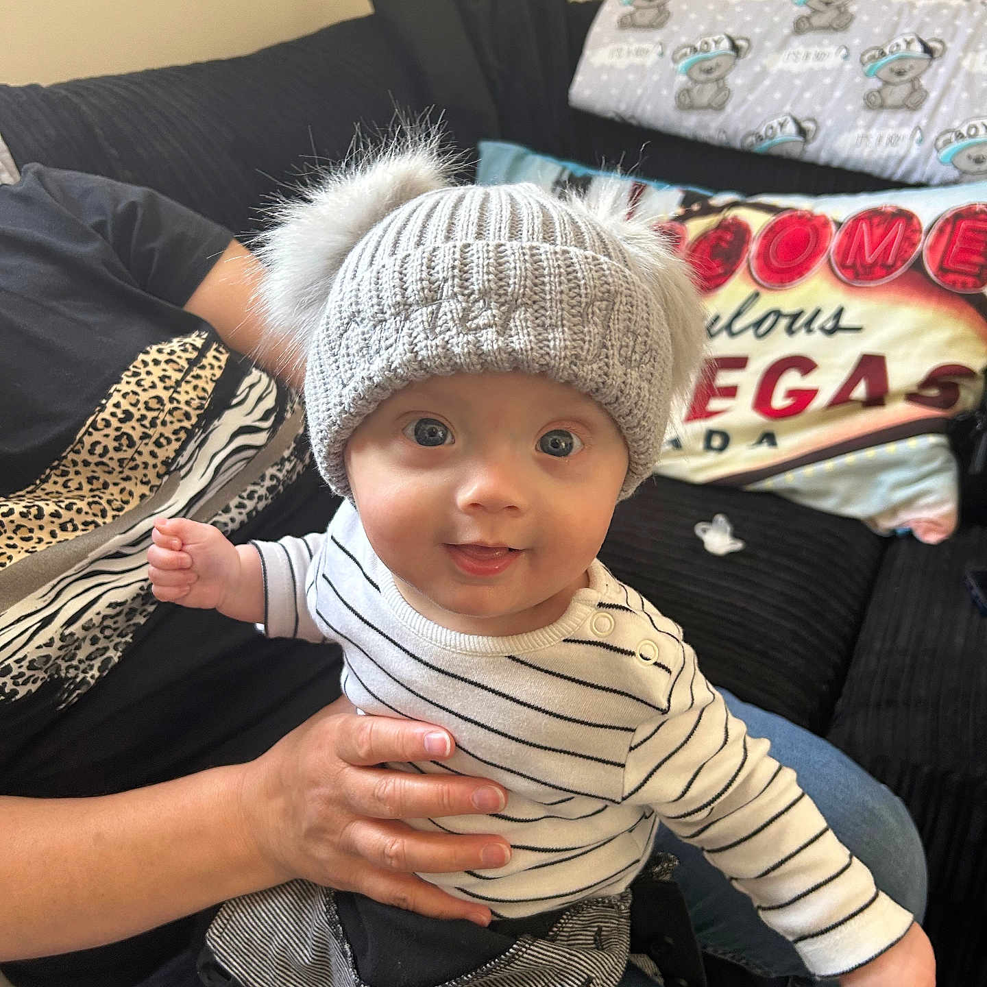 Carter-Jae is registered to the contest to win money with this photo: adult_hand, baby, black_pants, blue_eyes, child, couch, cozy, cute, hand, happy, hat, indoor, jeans, living_room, person, pillow, pom_pom, portrait, smile, striped_shirt