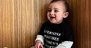 Asaiah is registered to the contest to win money with this photo: toddler, child, smiling, sitting, black_tshirt, checkered_pants, sneakers, wooden_bench, happy, indoor, cute, fashion, casual_clothing, short_hair, orange_socks, portrait, one_person, young_child, funny_tshirt, playful