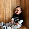 toddler, child, smiling, sitting, black_tshirt, checkered_pants, sneakers, wooden_bench, happy, indoor, cute, fashion, casual_clothing, short_hair, orange_socks, portrait, one_person, young_child, funny_tshirt, playful