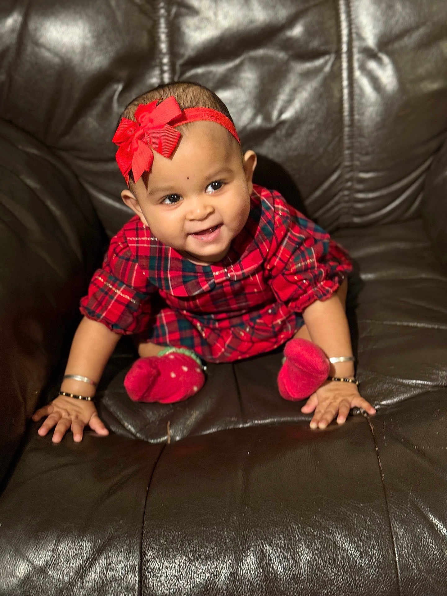 Jisha joined the competition — help win amazing prizes! baby, child, red_bow, headband, plaid_dress, red_socks, bracelets, smiling, leather_couch, brown_couch, happy, cute, portrait, sitting, infant, indoors, person, furniture, young_child, adorable
