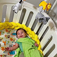 Patrick joined the competition — help win amazing prizes! baby, crib, blanket, bedding, stuffed_animals, mobile, yellow, green, smiling, child, infant, cute, playful, indoor, furniture, happy, animal_print, toddler, resting, bedroom