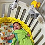 baby, crib, blanket, bedding, stuffed_animals, mobile, yellow, green, smiling, child, infant, cute, playful, indoor, furniture, happy, animal_print, toddler, resting, bedroom