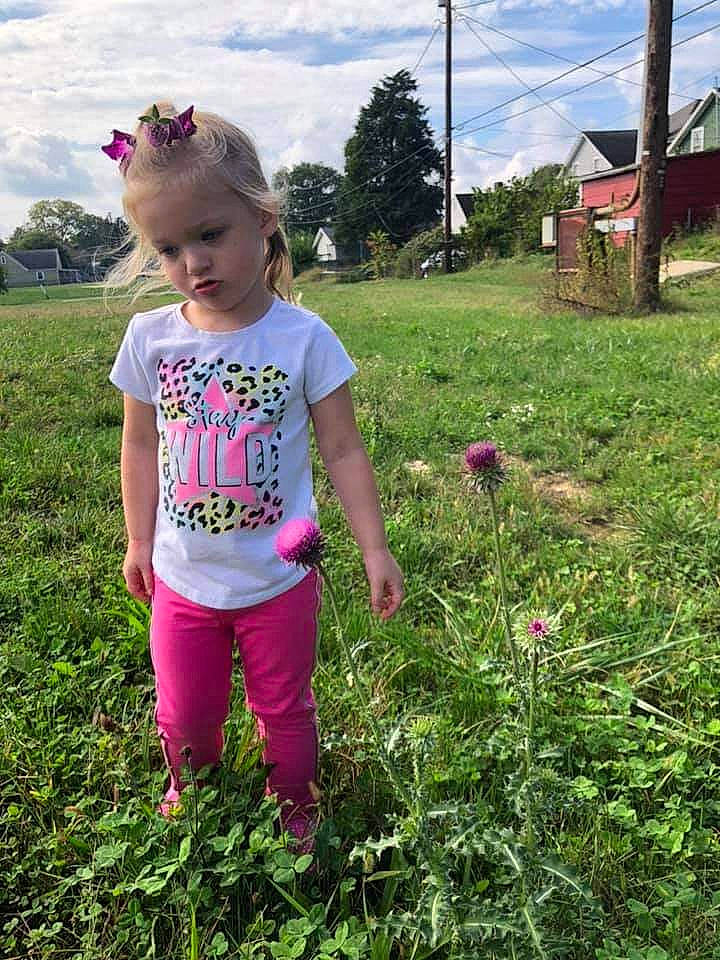 Addilynne is registered to the contest to win money with this photo: baby_toddler_clothing, botany, cloud, flower, grass, grassland, groundcover, happy, magenta, meadow, natural_environment, nature, people_in_nature, person, plant, rural_area, sky, sleeve, t_shirt, terrestrial_plant