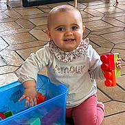 Victoire a rejoint le concours — aidez-le/la à gagner de superbes lots ! baby, child, smiling, playing, toys, building_blocks, floor, indoor, container, colorful, clothing, person, happy, cute, face, head, kneeling, toy_blocks, pink_pants, white_shirt