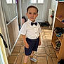 child, boy, smiling, indoor, hallway, shirt, bow_tie, shorts, socks, floor_tiles, mirror, shoes, cabinet, radiator, door, light, clothing, person, portrait, standing