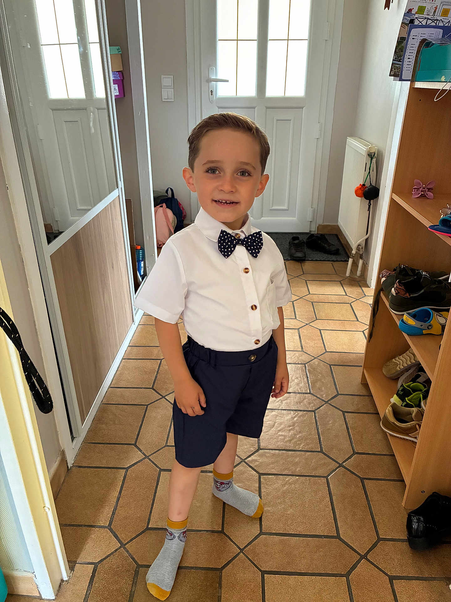 Arthur a rejoint le concours — aidez-le/la à gagner de superbes lots ! child, boy, smiling, indoor, hallway, shirt, bow_tie, shorts, socks, floor_tiles, mirror, shoes, cabinet, radiator, door, light, clothing, person, portrait, standing