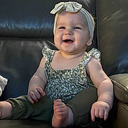 Evelyn is registered to the contest to win money with this photo: baby, smiling, headband, green_pants, sitting, leather_couch, happy, child, infant, cute, portrait, indoors, young_child, clothing, foot, hand, face, person, furniture, head