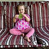 barefoot, blonde_hair, child, couch, daylight, face, floral_shirt, home, leaf, nature, outdoor, pink_pants, playful, portrait, sitting, smiling, striped_cushion, toddler, window, wooden_floor